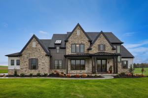 exterior of custom home with beige and brown stone siding, light and brown accent siding, dark roof, covered front porch, french doors, curved paved front walkway and flowerbeds